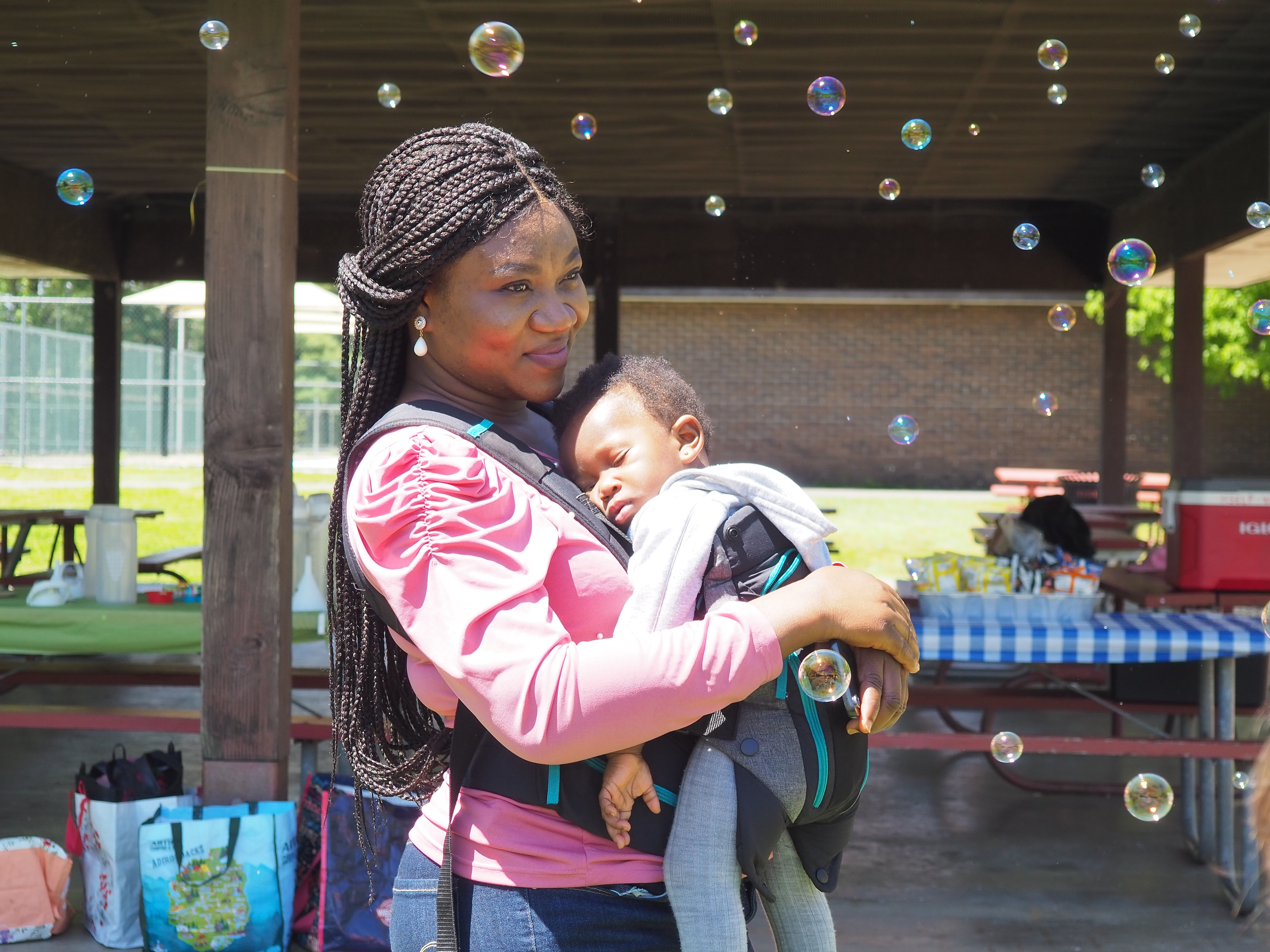 A Black mother with long braids smiles and hugs her sleeping child to her chest; they are surrounded by floating bubbles